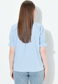 Top azzurro chiaro a maniche corte con dettagli arricciati sulle maniche. Tessuto morbido, vestibilità comoda e orlo curvo sul retro. Indossato con jeans chiari.