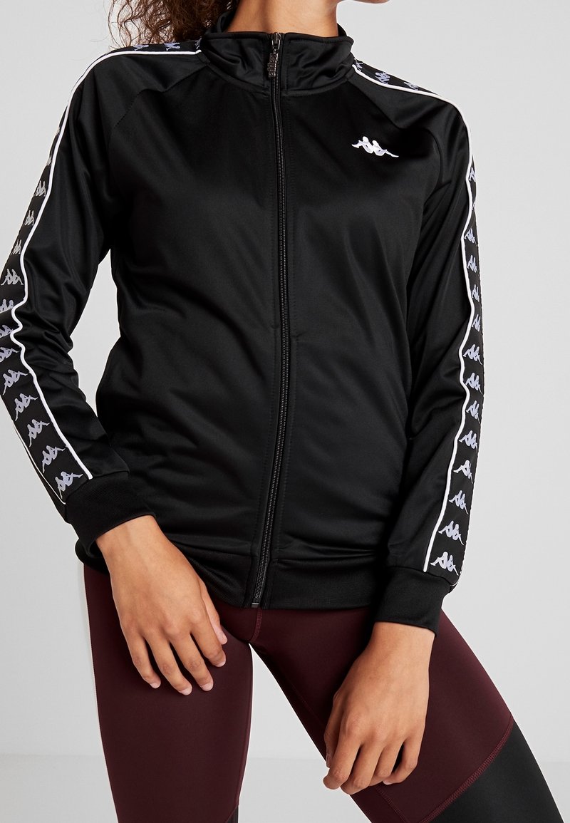 Personne portant une veste noire zippée avec des bandes blanches avec logo sur les manches et des leggings bordeaux, mains reposant sur les cuisses.