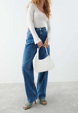 Top bianco a maniche lunghe con scollatura rotonda, abbinato a jeans blu a vita alta e gamba larga. Tiene una piccola borsa a tracolla in pelle bianca. Sandali verdi con tacco.