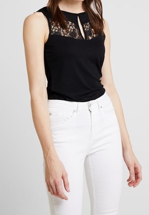 Czarny top bez rękawów z koronkowym karczkiem i dekoltem w kształcie łezki, zestawiony z białymi dżinsami z wysokim stanem na stojącej kobiecie.