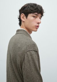 Hnědý vlněný svetr s límcem, s volným střihem a jemnou texturou, pohled ze strany na neutrálním pozadí.