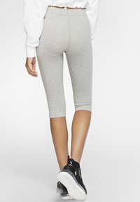 Šedé bavlněné legíny capri dosahují k polovině lýtek a mají přiléhavý střih. Jsou kombinovány s černými sportovními botami a bílým topem, viděny zezadu.