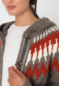 Dzianinowy kardigan w kolorach brązowym, pomarańczowym i białym z kapturem. Charakteryzuje się teksturowanym wzorem i zapięciem na zatrzaski z kontrastującą wstawką.