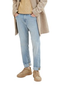 Blugi din denim bleu deschis cu manșete răsucite, asociați cu adidași bej. O haină bej și un pulover texturat galben muștar completează ținuta.