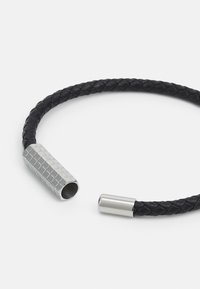 Bracciale in pelle intrecciata nera con hardware in argento testurizzato e estremità cilindriche. Gli accenti metallici presentano un motivo geometrico.