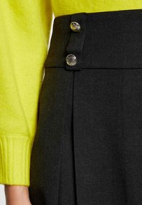 Pantaloni neri a vita alta caratterizzati da due bottoni argentati in vita, con un design plissettato, abbinati a un maglione in maglia gialla brillante.