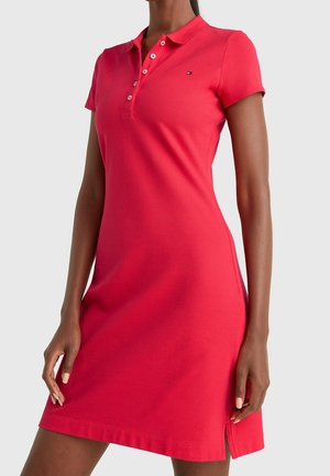 Femme portant une robe polo rouge moulante à manches courtes avec des boutons et une petite fente latérale, debout devant un fond uni.