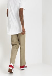 Osoba v béžových kalhotách, bílém tričku a červených kostičkovaných teniskách, která ukazuje tetování na své levé předloktí, kráčí proti jednoduchému bílému pozadí.