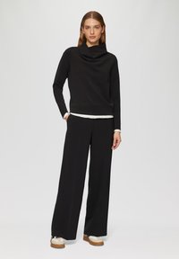 Schwarzes Oberteil mit Kapuze, kombiniert mit weiten schwarzen Hosen. Das Outfit hat eine gestufte weiße Unterschicht und lässige Sneakers mit einer strukturierten Sohle.