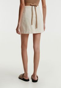 Sahnemarfarbener Baumwoll-Minirock mit gebundenem Bund, flacher Vorderseite und einfachen Stichakzenten. Kombiniert mit braunen Slipper-Sandalen.