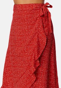 Falda corta roja con patrón floral blanco, dobladillo con volantes y detalle de lazo lateral. Hecha de una tela ligera, con superficie texturizada.
