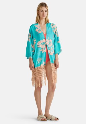 Kimono turquoise avec des motifs floraux roses, doté d'un ourlet en franges au crochet, superposé sur un maillot de bain corail vif.