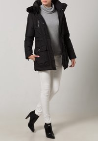 Giacca nera imbottita con cappuccio in pelliccia sintetica, due tasche frontali e chiusura con zip, indossata sopra un maglione grigio e abbinata a jeans skinny bianchi.
