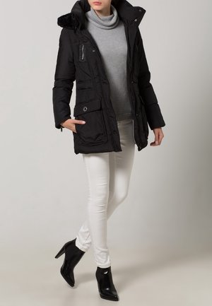 Sort polstret jakke med faux fur hætte, to forlommer og lynlås, båret over en grå sweater og parret med hvide skinny jeans.