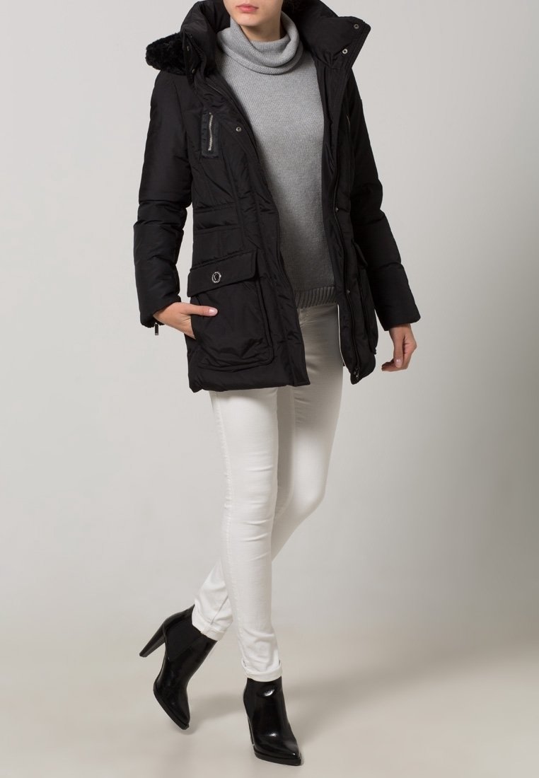 Giacca nera imbottita con cappuccio in pelliccia sintetica, due tasche frontali e chiusura con zip, indossata sopra un maglione grigio e abbinata a jeans skinny bianchi.