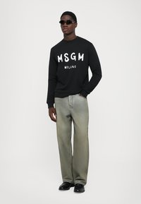 Schwarzer Hoodie mit weißem "MSGM MILANO"-Text, kombiniert mit hellgrauen Jeans. Das Model trägt dunkle Sonnenbrillen und schwarze Schuhe.