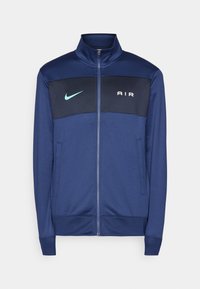 Marinblå zip-up jacka med hög krage, som har en svart bröstpanel, vit "AIR"-text och en turkos Nike-swoosh. Slät tyg med sidofickor.