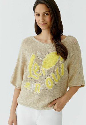 Femme aux longs cheveux foncés portant un haut en tricot beige avec un motif citron jaune et le texte « lemon oui », debout avec une main dans la poche d'un pantalon blanc.