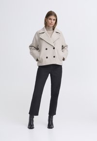 Beige double-breasted wool coat with black buttons, side pockets, and wide lapels. Paired with black straight-leg jeans and ankle boots.