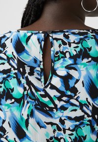 Blouse multicolore avec un motif floral dans des tons de bleu, vert et noir. Le dos présente une fermeture à bouton et une ouverture en forme de clé. Tissu texturé.