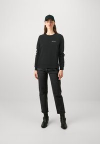 Felpa nera con colletto rotondo e maniche lunghe, abbinata a jeans neri e stivaletti. Un berretto nero completa l'outfit.