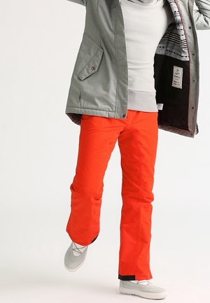 Orange waterproof ski pants with a straight leg design, paired with a gray jacket and white footwear, showcasing a layered fabric texture.