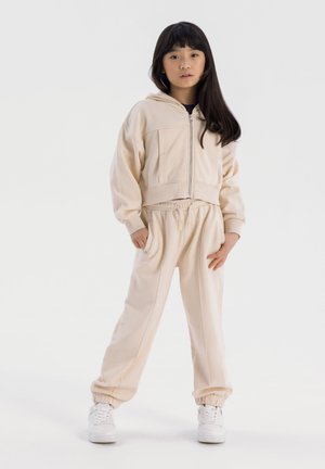 Ensemble de vêtements de détente beige comprenant un hoodie cropped à zip avec poches avant et un pantalon à taille élastique avec des poignets fuselés. Porté avec des baskets blanches.