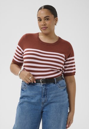 Žena s tmavými vlasmi, oblečená v hnedo-ružovom pruhovanom tričku s krátkym rukávom, modrých džínsoch a čiernom opasku, nosiaca prstene a náramky, stojaca pred jednoduchým pozadím.