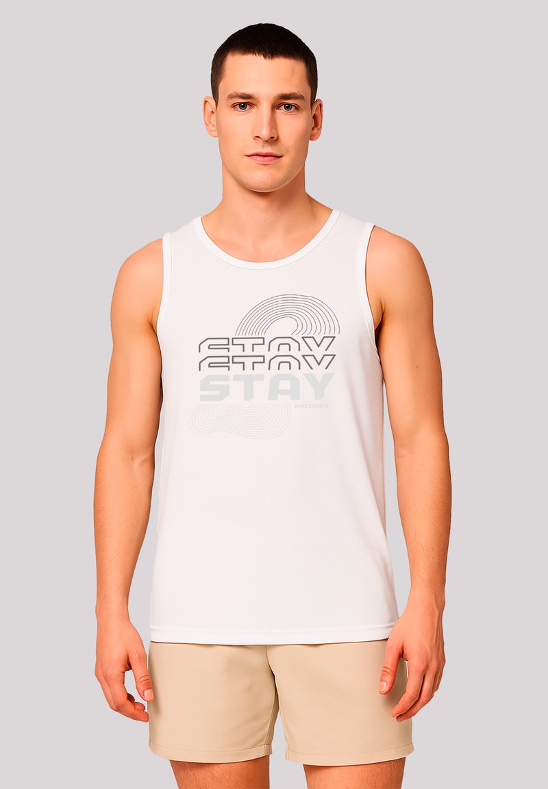 Mann in einem weißen ärmellosen T-Shirt mit dem Aufdruck "STAY" und beigen Shorts, der vor einem einfarbigen hellgrauen Hintergrund steht.