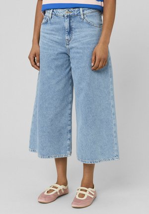 Personne portant un jean large court bleu clair et des ballerines roses avec des lanières blanches, debout devant un fond uni.