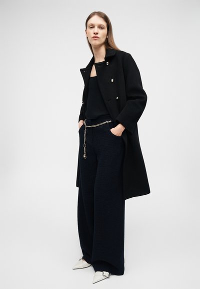 Γυναίκα που στέκεται με τα χέρια στις τσέπες, φοράει μαύρο παλτό με ασημένια κουμπιά, φαρδιά μαύρα παντελόνια, ζώνη με αλυσίδα και λευκά μυτερά παπούτσια.