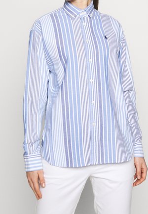 Kobieta w niebiesko-białej koszuli z pionowymi paskami zapinanej na guziki z długimi rękawami i białych spodniach, stojąca na jednolitym tle.