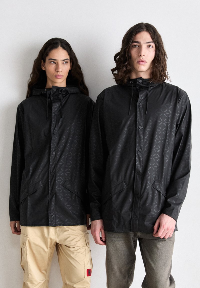 Vestes noires imperméables avec un motif géométrique, boutons à l'avant et poches latérales. Les modèles portent des pantalons de teintes neutres, se tenant devant un fond uni.