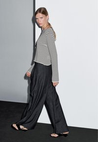 Gestreept zwart-wit langemarige top, gecombineerd met donkere broek met wijde pijpen. De textuur lijkt glad, met minimale accenten en neutrale tinten.