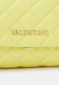 Quiltad gul läderväska med en guld "Valentino"-logotyp och en guldspänne, som framhäver en slät textur och geometriskt mönster.