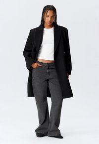Woman with braided hair wearing a black oversized coat, white cropped top, wide-leg gray jeans, and black shoes, standing with one hand in pocket.