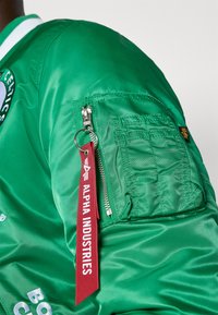 Jachetă bomber verde cu un finisaj lucios, având un buzunar cu fermoar pe mânecă și o etichetă roșie inscripționată "ALPHA INDUSTRIES".