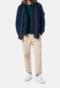 Marinblå puffjacka, grön polo skjorta, krämfärgade byxor och vita sneakers. Halsduk med ett texturerat mönster hängande runt halsen.