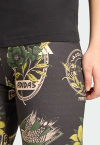 T-shirt preta e leggings estampadas com desenhos de flores e pássaros, principalmente em verde, amarelo e cinza, com um detalhe do logótipo da Adidas.