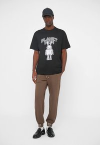 Zwart katoenen T-shirt met een witte robotafbeelding en de tekst "PLANET MCM", gecombineerd met bruine joggingbroek en zwarte schoenen.