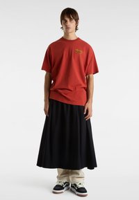 Rød bomulds T-shirt med "Vans" logo, parret med en lang sort nederdel. Modellen bærer sorte sneakers med hvide og brune accenter.