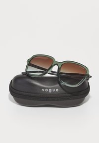 Ochelari de soare verzi, supradimensionați, cu lentile maro degradé, așezați pe un etui negru cu fermoar, pe care este imprimat cu alb "vogue".