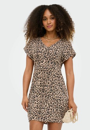 Femme aux cheveux bouclés portant une robe à manches courtes imprimé léopard et tenant un petit sac à main beige avec un détail chaîne dorée.