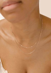 Dos delicados collares de plata de diferentes grosores caen suavemente alrededor del cuello, descansando contra una clavícula lisa y desnuda.