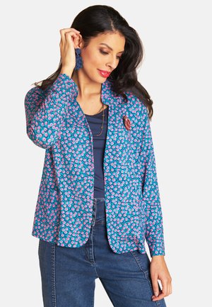 Woman adjusting blue floral earrings wearing blue patterned blazer, navy top, cross necklace, and blue jeans against white background.
