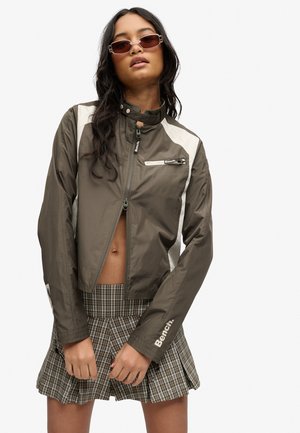 Frau trägt braun-weiße Bench-Zipjacke, karierter Faltenrock und rechteckige Sonnenbrille, posiert mit Händen am Rocksaum.