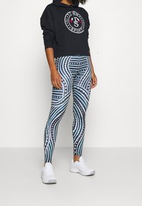 Leggings sportivi con un motivo a onde nero, blu e bianco, con accenti a forma di stella. Abbinati a un hoodie accorciato nero e a scarpe da ginnastica bianche.