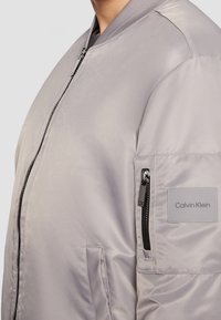 Giacca bomber grigio chiaro con una finitura liscia e lucida; presenta una chiusura con zip, colletto a coste e una tasca sulla manica sinistra con un'etichetta.