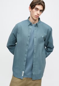 Svijetloplava košulja na dugih rukava od mekog pamuka, s klasičnim ovratnikom i malim logotipom na prsima, složena preko tirkizne majice.