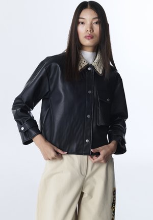 Woman poses wearing black leather jacket with beige textured collar and beige pants with floral embroidery on side against white background.